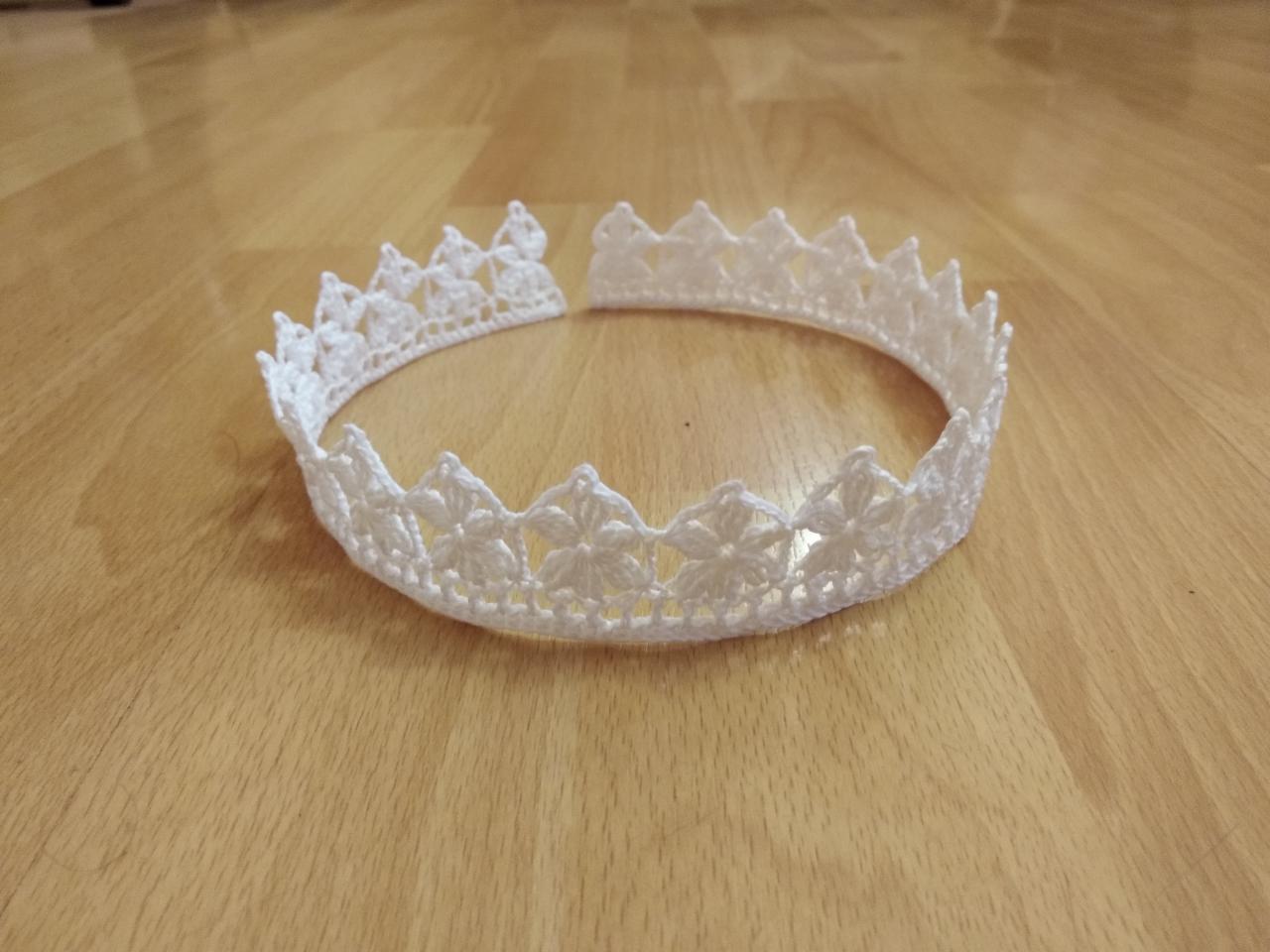 Biała, koronkowa, ręcznie robiona na szydełku korona z motywem kwiatowym, leżąca na drewnianej podłodze.