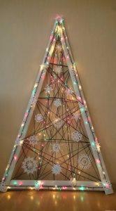 Ręcznie robiona choinka z drewnianej drabiny, ozdobiona białymi, ażurowymi płatkami śniegu i kolorowymi lampkami, na tle beżowej ściany.