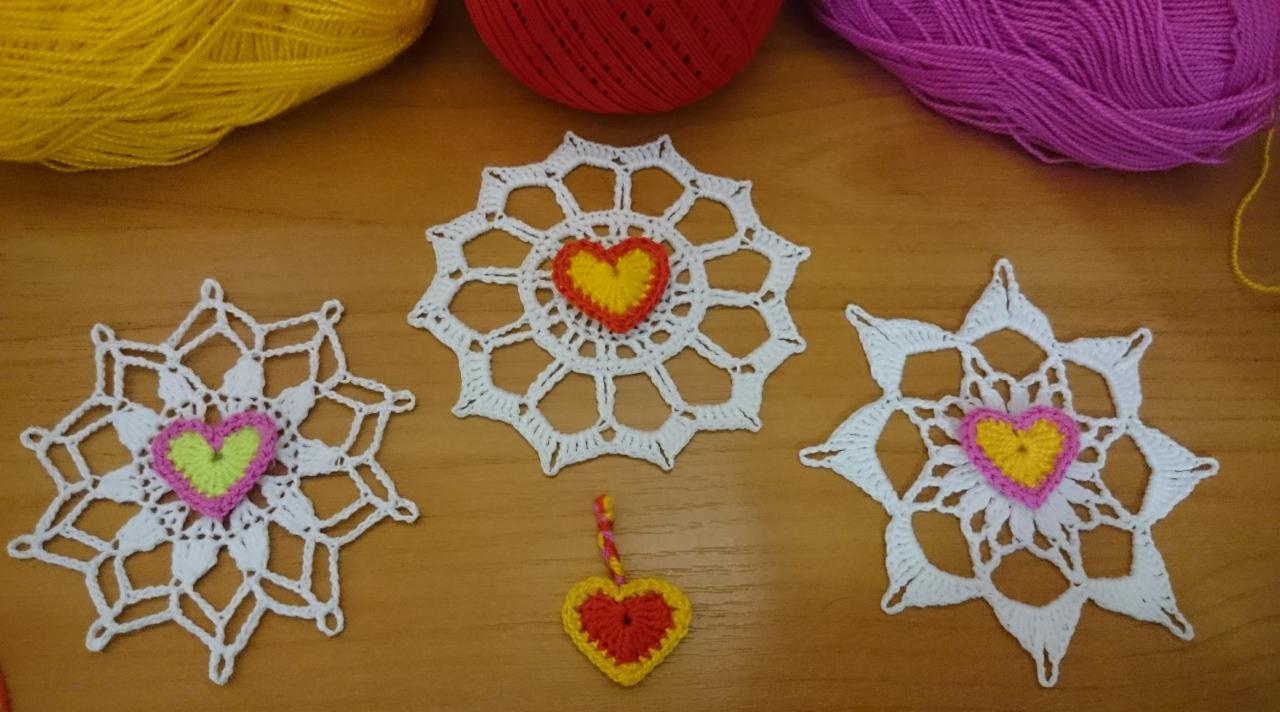 Trzy białe, ażurowe serwetki z szydełka z kolorowymi sercami w środku (żółte, różowe, zielone) oraz mała zawieszka w kształcie serca na drewnianym stole, obok leżą kolorowe kłębki wełny.