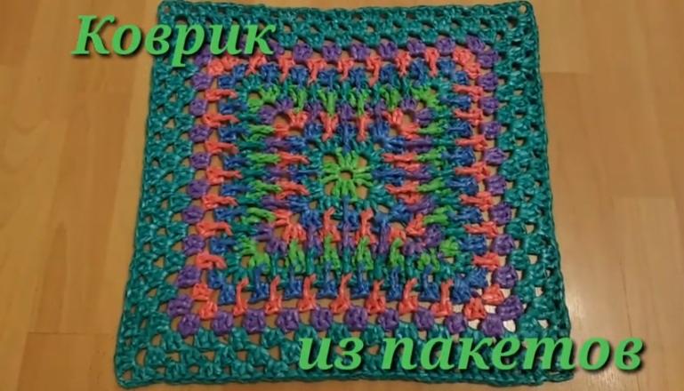 Kolorowy, kwadratowy dywanik wykonany szydełkiem z recyklingowych toreb foliowych, leżący na drewnianej podłodze.