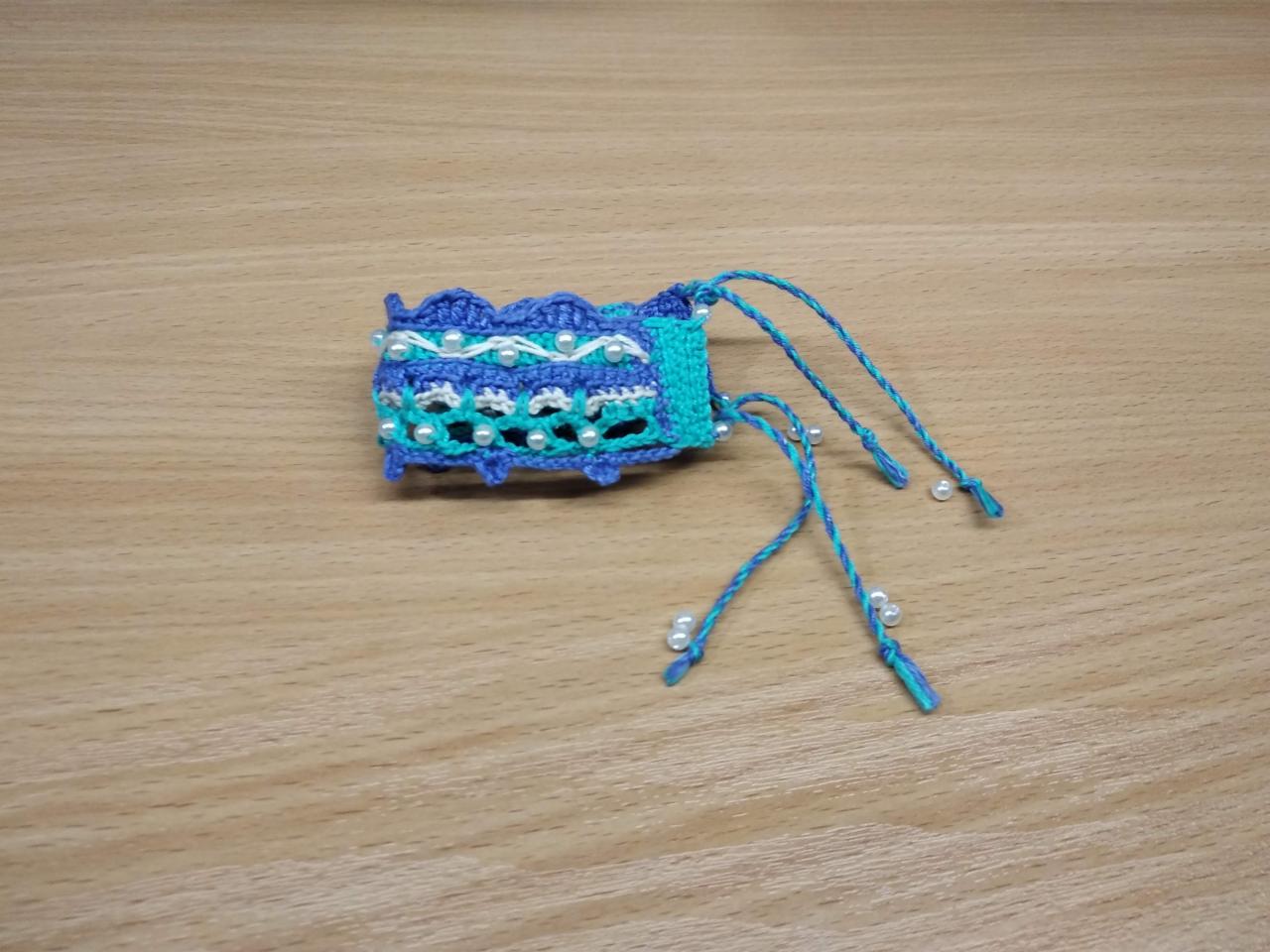 Ręcznie robiona, niebiesko-turkusowa bransoletka na szydełku, ozdobiona białymi perłami, leżąca na drewnianym blacie.