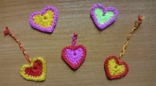 Ręcznie robione, kolorowe serduszka z włóczki, dwa z zawieszkami z plecionego sznurka, ułożone na drewnianej powierzchni. Jedno serce czerwono-żółte z zawieszką, drugie różowo-żółte, trzecie...