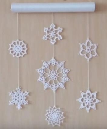 Ręcznie robiona, biała, ażurowa dekoracja ścienna w kształcie śnieżynek o różnych wzorach, zawieszona na drewnianym drążku na tle beżowej ściany.