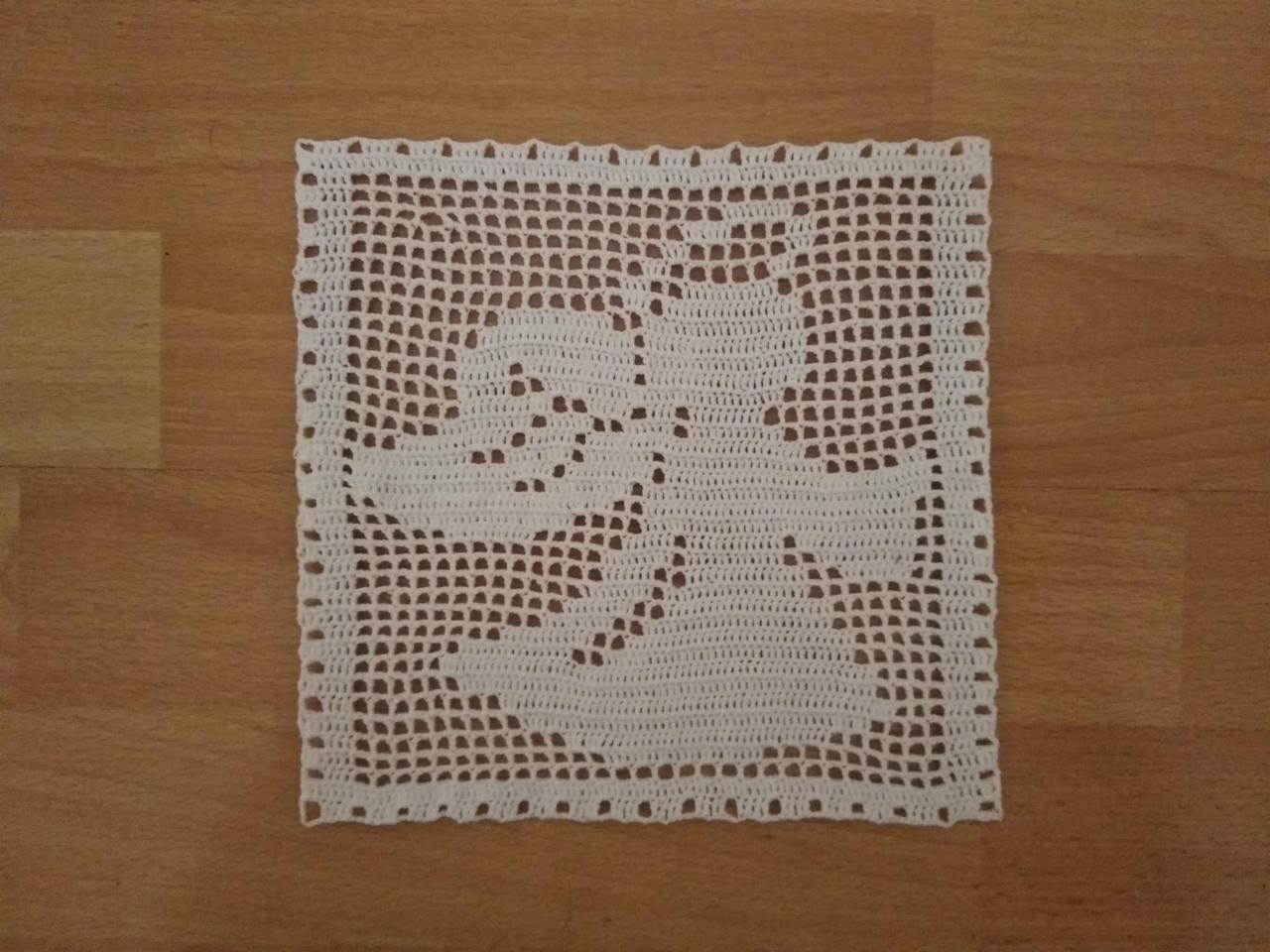 Kwadratowa, ręcznie robiona serwetka z ażurowym wzorem dwóch gołębi, leżąca na drewnianym stole.