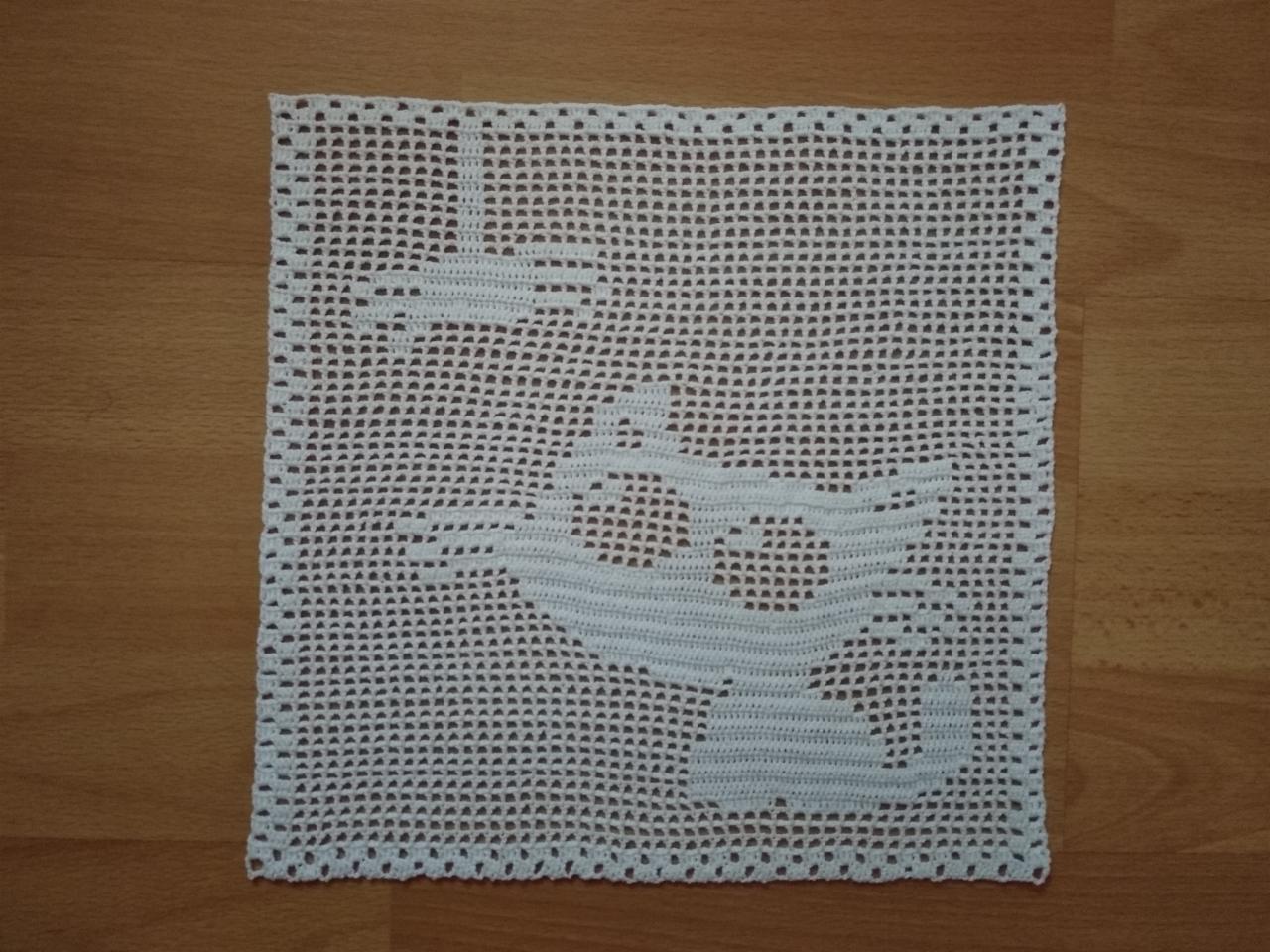 Kwadratowa, biała serwetka z ażurowym wzorem anioła i gwiazdy, leżąca na drewnianym blacie.