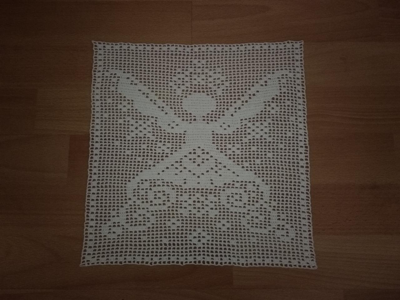 Biała, ręcznie robiona, kwadratowa serwetka z ażurowym wzorem, leżąca na drewnianej podłodze.