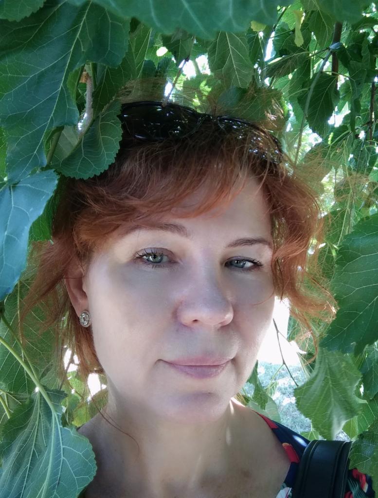 Portret kobiety z rudymi włosami, w zieleni liści, z okularami na głowie i kolczykami w uszach, z delikatnym uśmiechem.