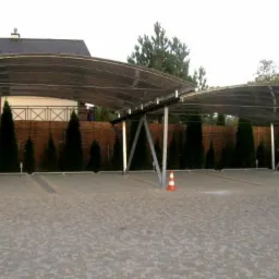 Dwa zadaszone miejsca parkingowe z przezroczystym dachem i metalowymi słupami na posesji z ogrodzeniem z drewnianych paneli i ozdobnymi drzewkami.