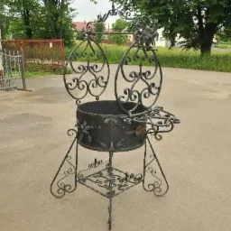 Kuty, ozdobny kwietnik z metalu, z dekoracyjnymi elementami roślinnymi, stojący na betonowym podłożu, w tle fragmenty ogrodzeń i zieleni.