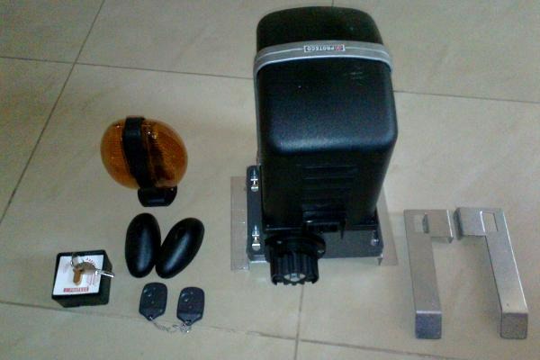 Zestaw do automatyzacji bramy przesuwnej: napęd z lampą ostrzegawczą, piloty, fotokomórki, kluczyk i elementy montażowe na kafelkowej podłodze.