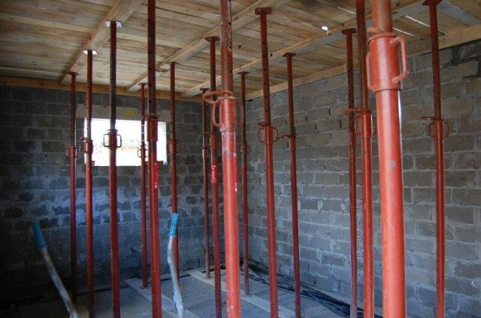 Wnętrze budynku w trakcie budowy, widoczne czerwone, metalowe podpory stropowe rozmieszczone równomiernie, podtrzymujące drewniany strop, z fragmentem okna w tle i surową, szarą ścianą z cegieł.