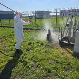Pracownik w kombinezonie ochronnym czyści element konstrukcyjny myjką ciśnieniową na zewnątrz, trawa z ż&oacute;łtymi kwiatami w tle, ogrodzenie i budynki w oddali.