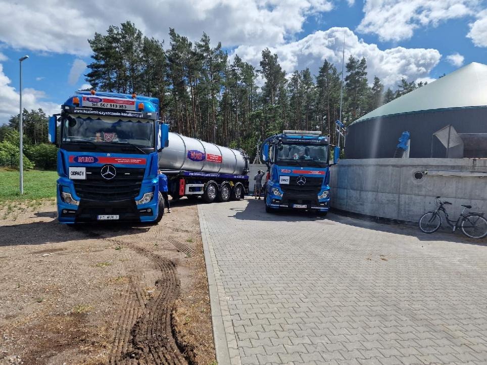 Dwa niebieskie ciągniki siodłowe z cysternami na parkingu z kostki, w tle las i biogazownia z rowerem opartym o betonowy mur.