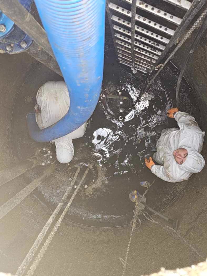 Wnętrze studni kanalizacyjnej z dwoma pracownikami w białych kombinezonach ochronnych i pomarańczowych rękawicach, niebieski wąż oraz drabina metalowa, widok z góry.