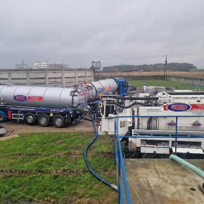 Dwa pojazdy asenizacyjne WUKO podłączone wężami podczas pracy na terenie oczyszczalni ścieków, widoczne zbiorniki, pompy i infrastruktura zakładu w tle.