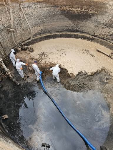 Czterech pracowników w białych kombinezonach, z użyciem węża i łopat, usuwa osad z betonowego zbiornika z grubymi ścianami, widok z góry.