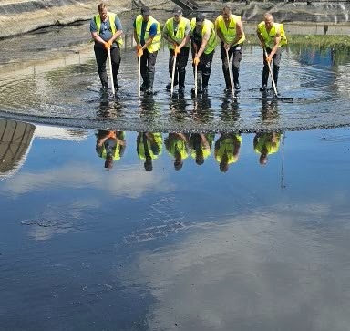 Sześciu pracowników w odblaskowych kamizelkach, stojących w rzędzie w zbiorniku z ciemną, prawdopodobnie przemysłową cieczą, używających narzędzi do mieszania lub czyszczenia, z odbiciem...