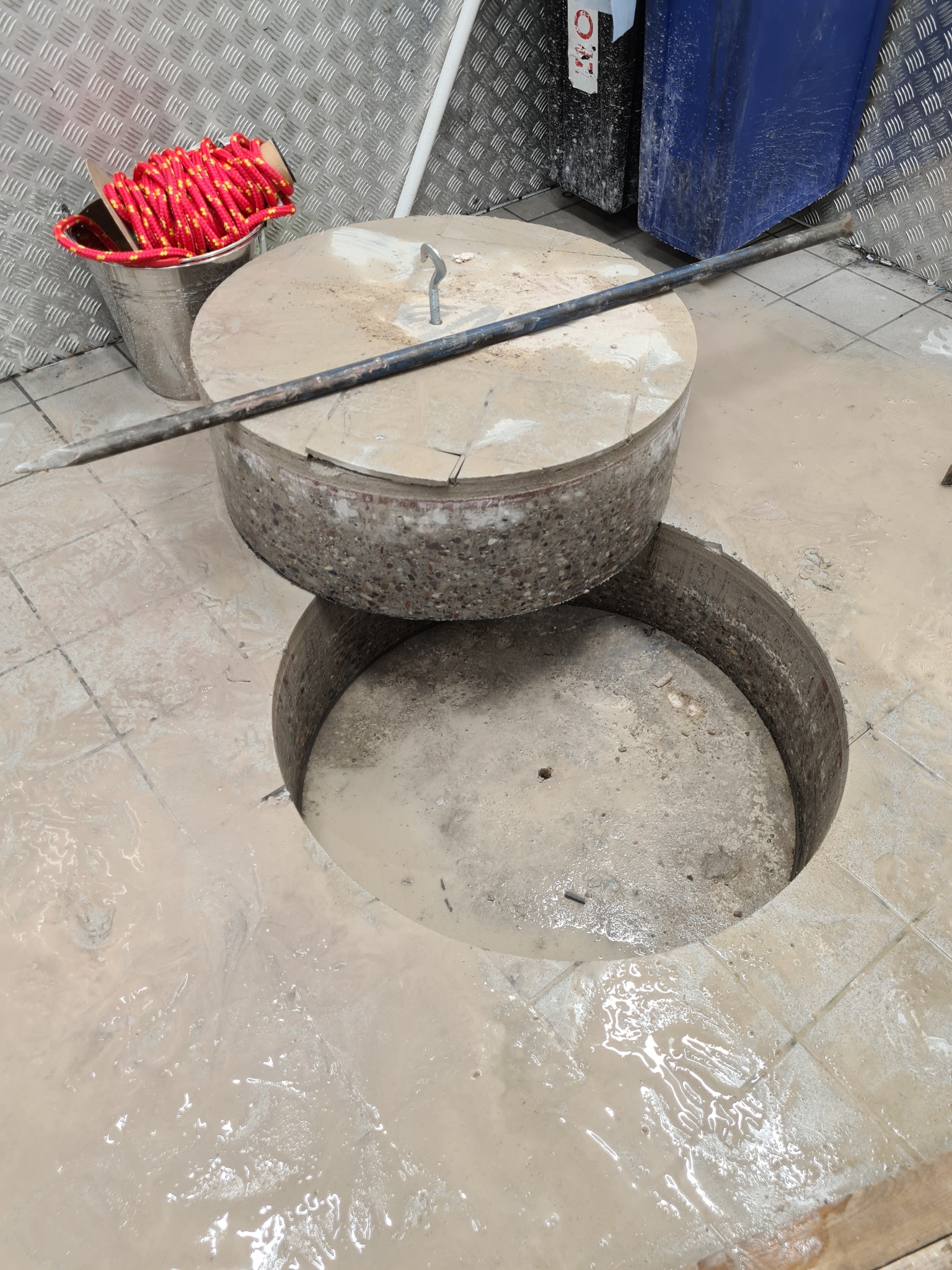 Otwór w betonowej posadzce z podniesionym, okrągłym fragmentem, obok wiadro z czerwoną liną i metalowy pręt, otoczenie zabrudzone.