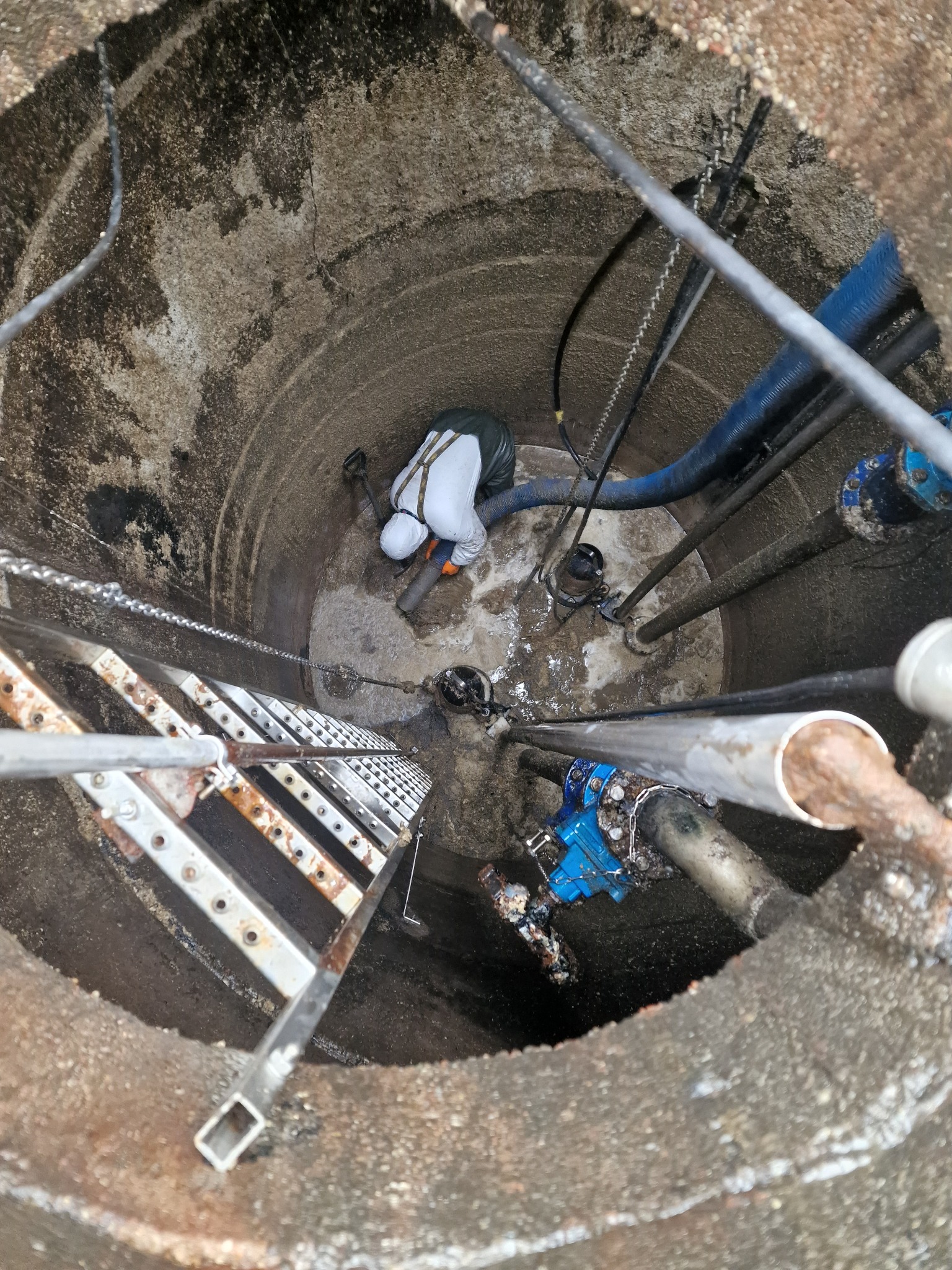 Pracownik w kombinezonie ochronnym wewnątrz betonowej studni z rurami i drabinką, podczas wykonywania prac konserwacyjnych.