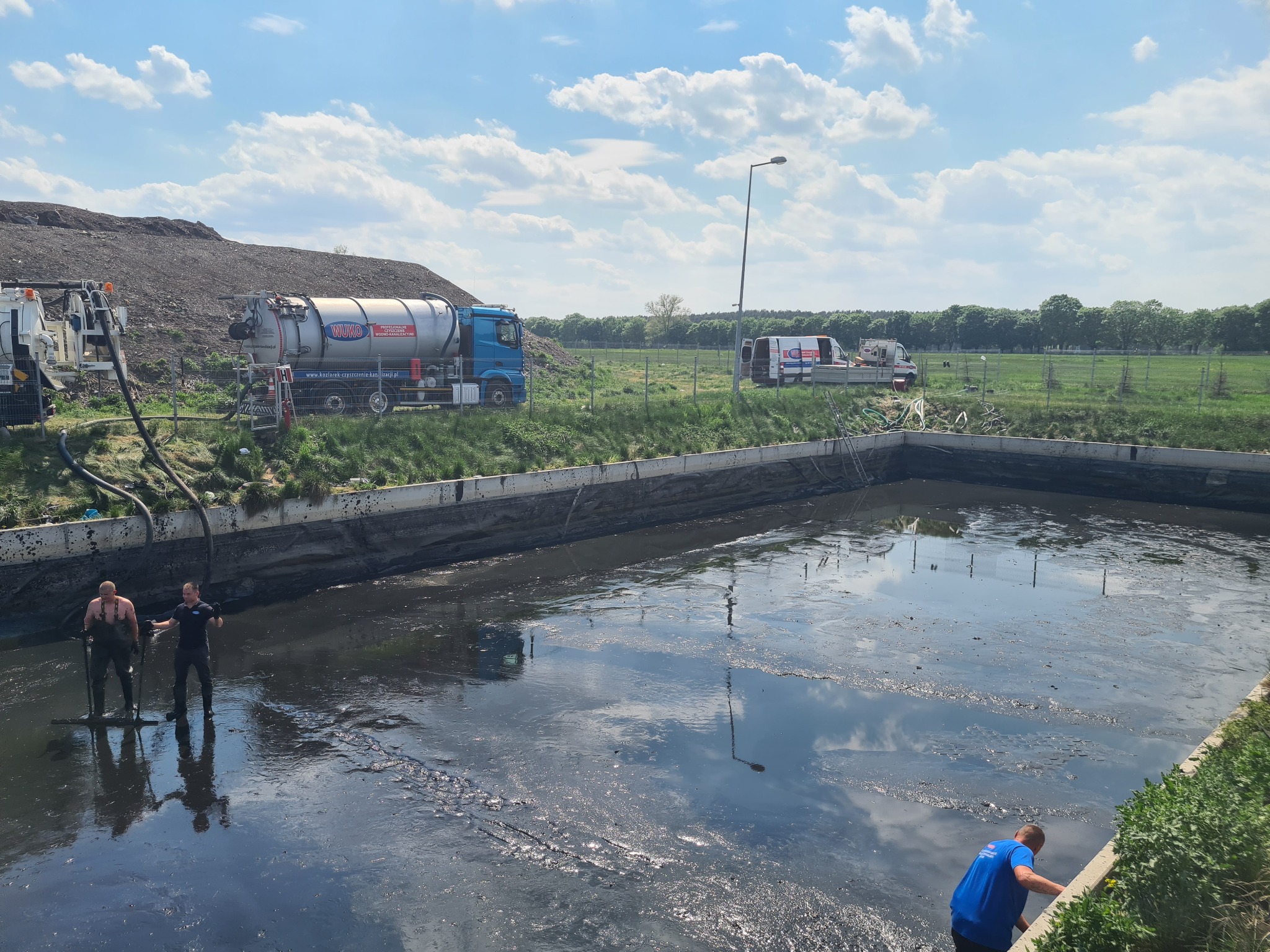 Dwóch pracowników w kombinezonach wchodzi do zbiornika z ciemną cieczą, w tle widoczne wozy asenizacyjne i składowisko odpadów pod błękitnym niebem.