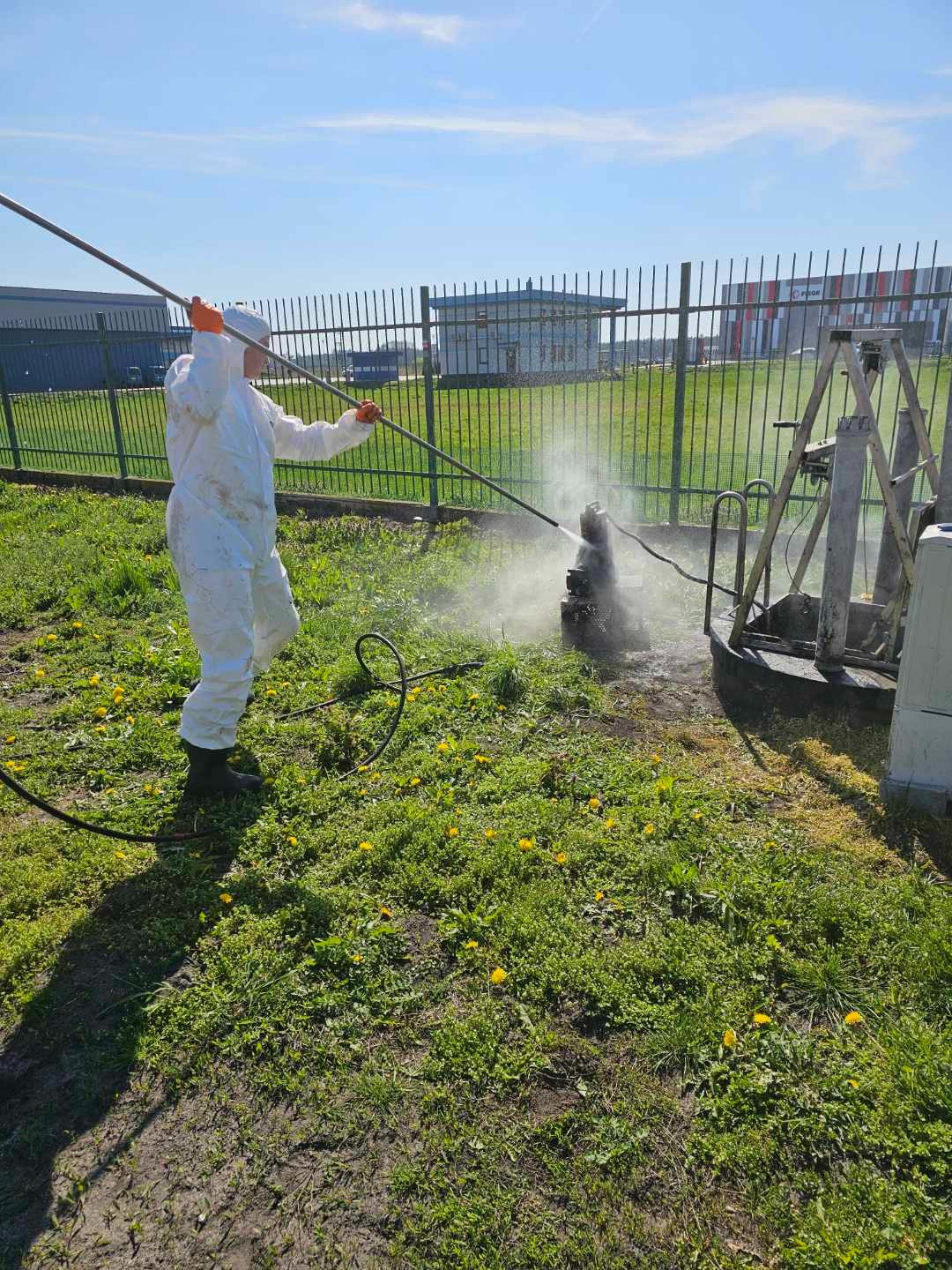 Pracownik w kombinezonie ochronnym czyści element konstrukcyjny myjką ciśnieniową na zewnątrz, trawa z żółtymi kwiatami w tle, ogrodzenie i budynki w oddali.