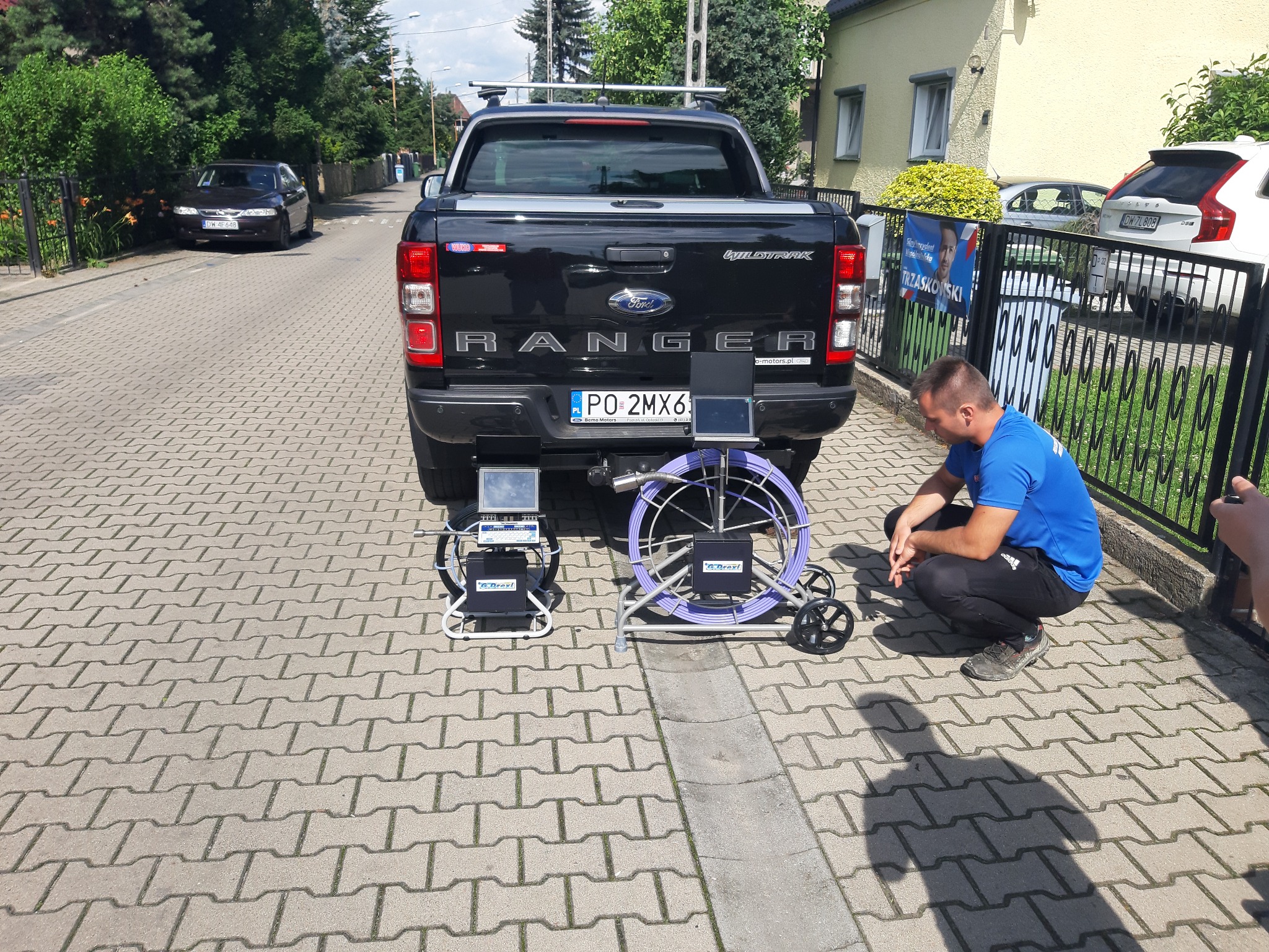 Czarny Ford Ranger z urządzeniem do inspekcji rur na chodniku przed domem, mężczyzna w niebieskiej koszulce kuca obok sprzętu.