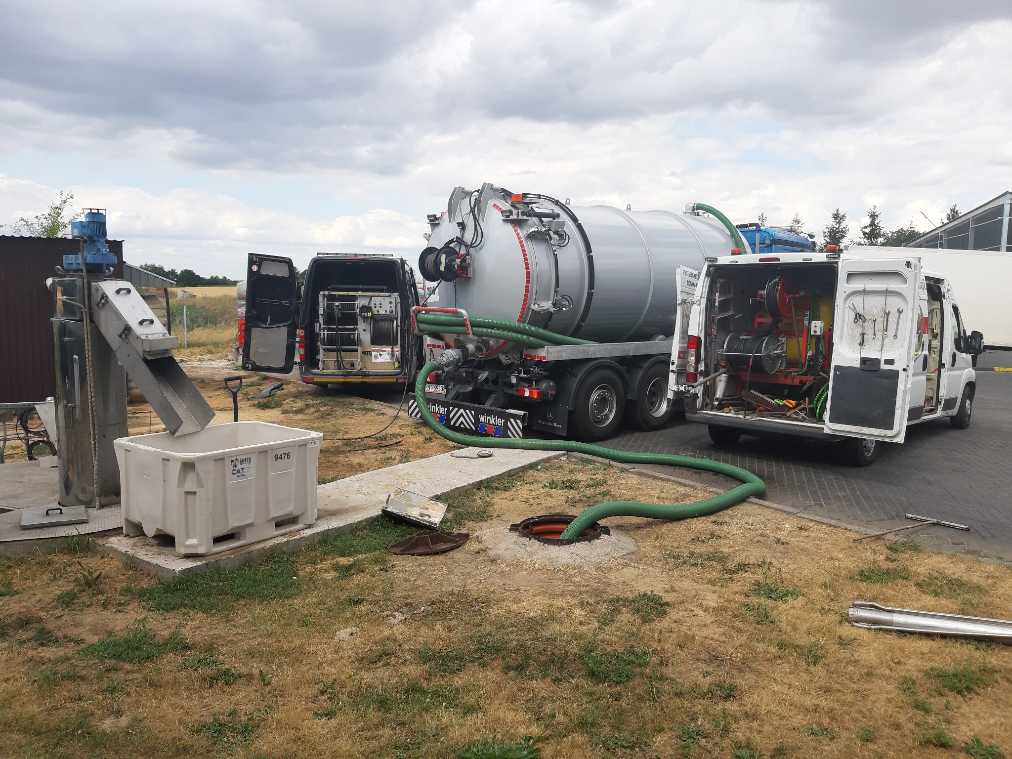 Ciężarówka asenizacyjna z otwartymi drzwiami, podłączona zielonym wężem do studzienki kanalizacyjnej, obok zaparkowany bus z wyposażeniem do czyszczenia kanalizacji i separator z pojemnikiem...
