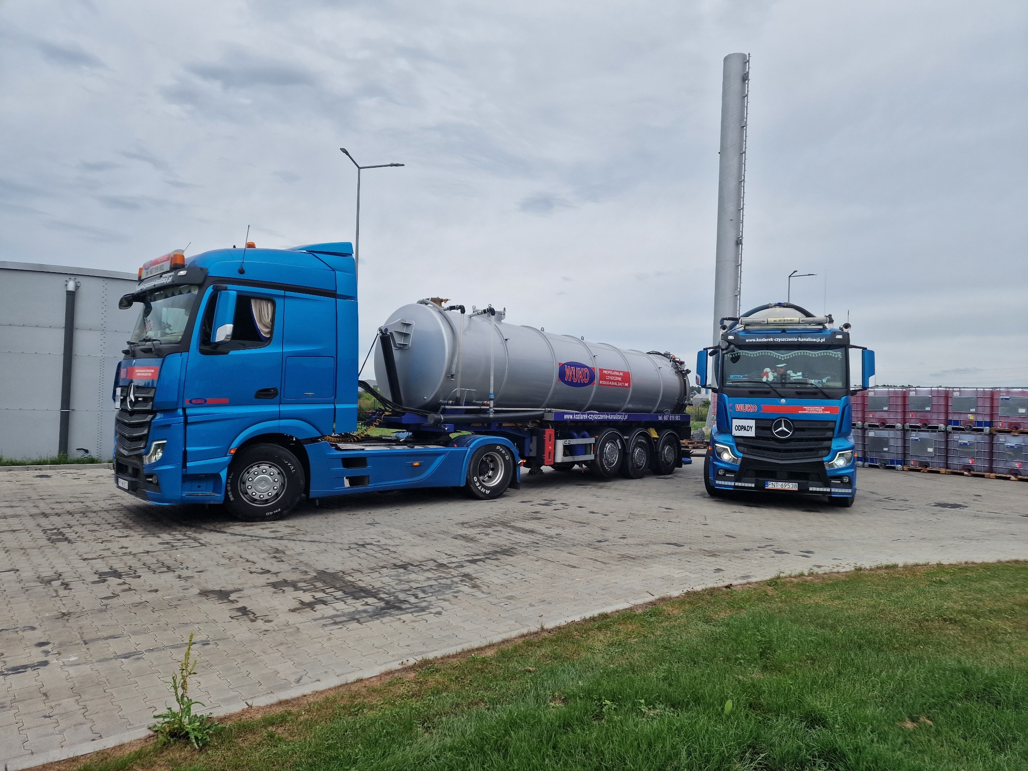 Dwa niebieskie ciągniki siodłowe Mercedes-Benz Actros z cysterną WUKO na naczepie, zaparkowane na placu z kostki brukowej w pochmurny dzień. Na cysternie widoczne logo firmy i numer telefonu. W tle...