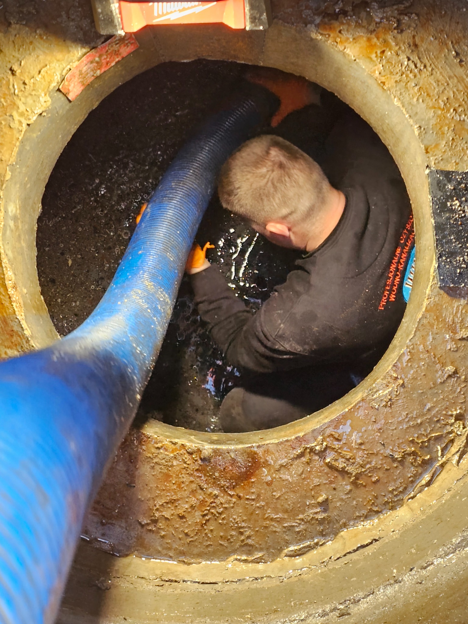 Pracownik w Dakowy Suche w trakcie czyszczenia studzienki kanalizacyjnej z użyciem niebieskiego węża ssącego, widoczny z góry w otworze studzienki.