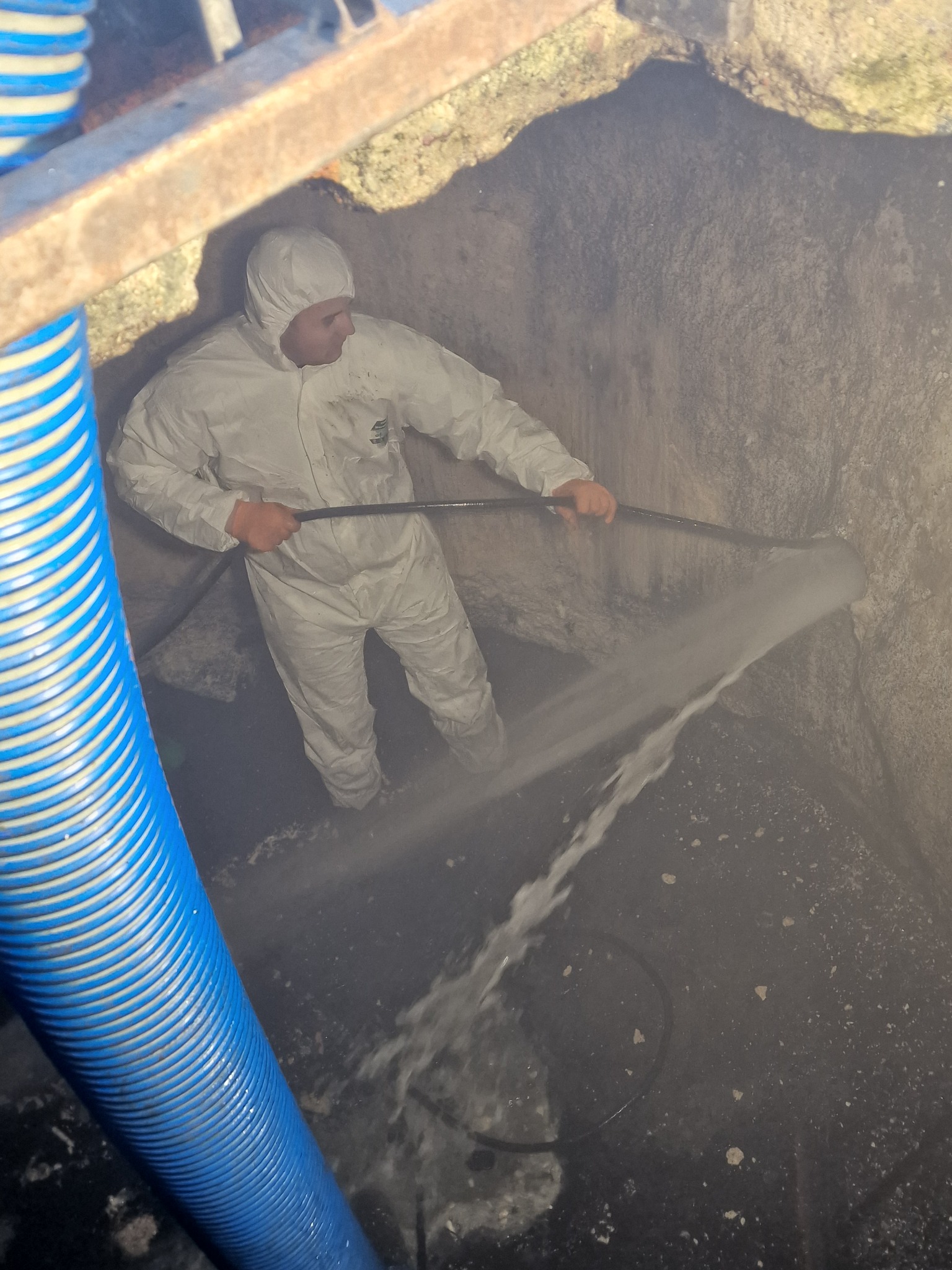 Pracownik w białym kombinezonie ochronnym czyści wnętrze betonowego szamba za pomocą węża ciśnieniowego, widoczny niebieski wąż doprowadzający wodę.