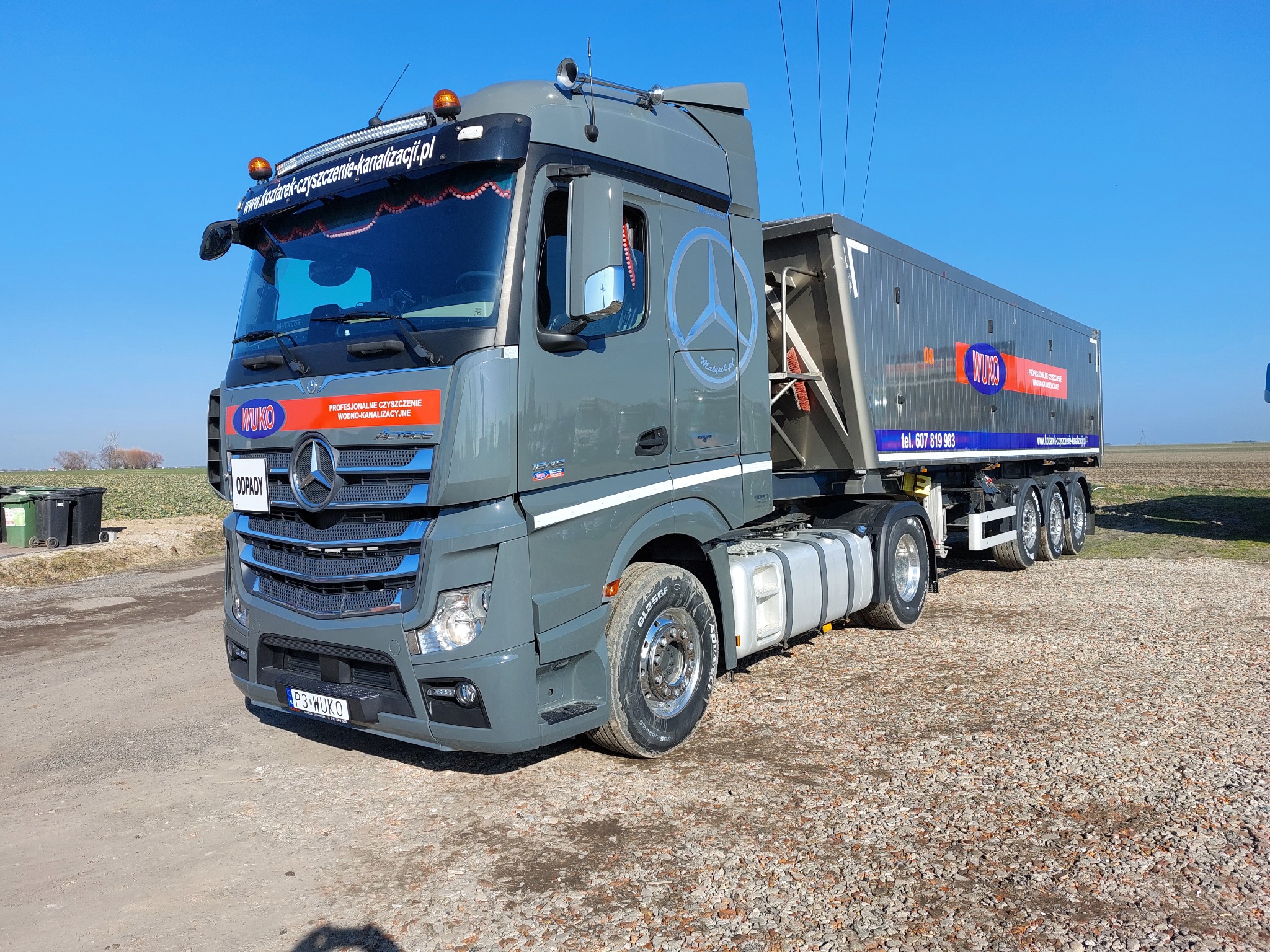 Szary ciągnik siodłowy Mercedes-Benz z naczepą, z logo firmy WUKO, na żwirowym podłożu, widoczne linie wysokiego napięcia i pole w tle, z naklejkami informującymi o czyszczeniu wodno-kanalizacyjnym.