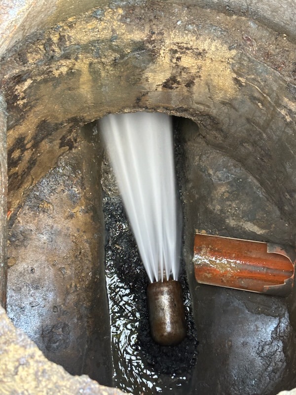 Dysza pod wysokim ciśnieniem usuwająca zator w studzience kanalizacyjnej, widoczne strumienie wody i fragment rury obok.