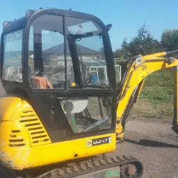 Żółta mini koparka JCB na gąsienicach, z łyżką lekko zanurzoną w ziemi, widoczna kabina operatora i otoczenie z zielenią oraz budynkiem w tle.