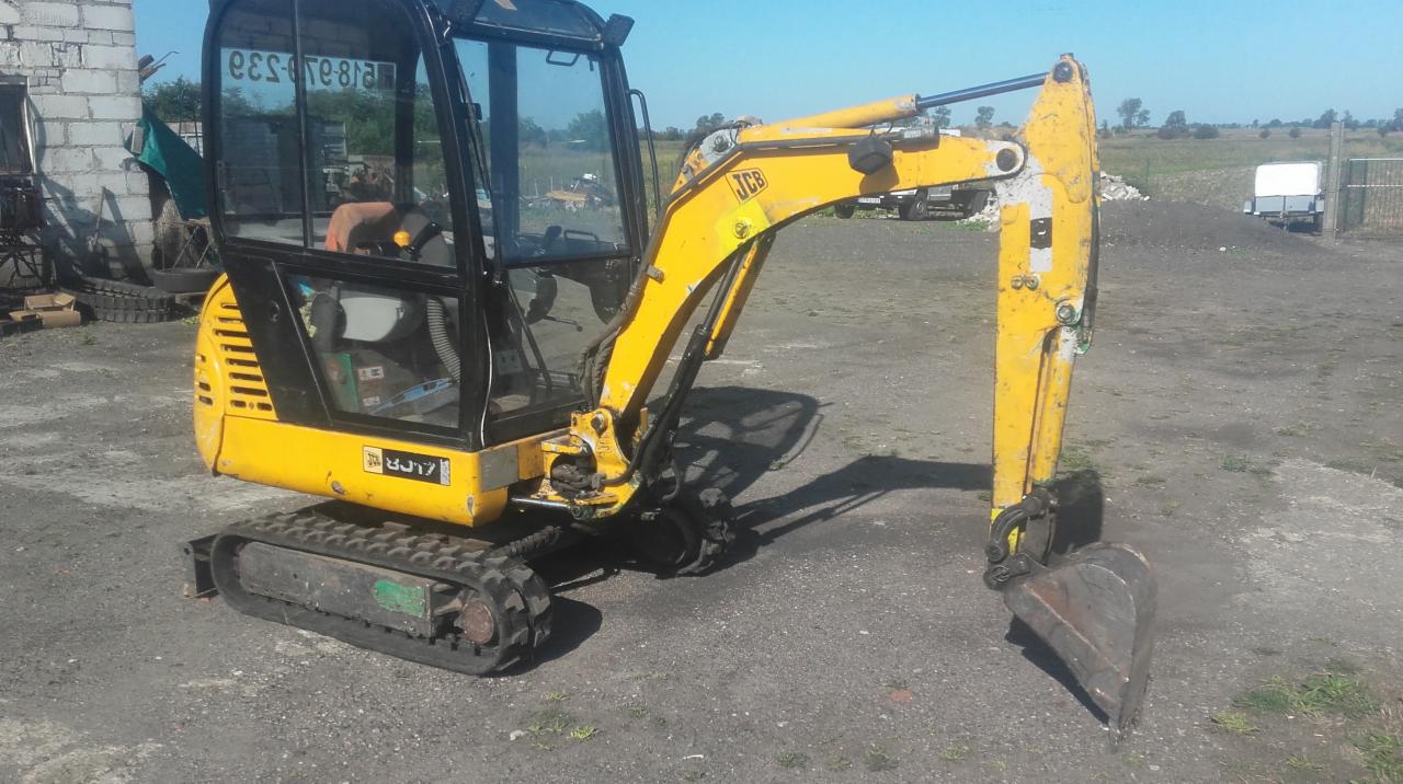 Żółta mini koparka JCB na placu budowy, widoczna kabina operatora i łyżka kopiąca, w tle budynek gospodarczy i zieleń.