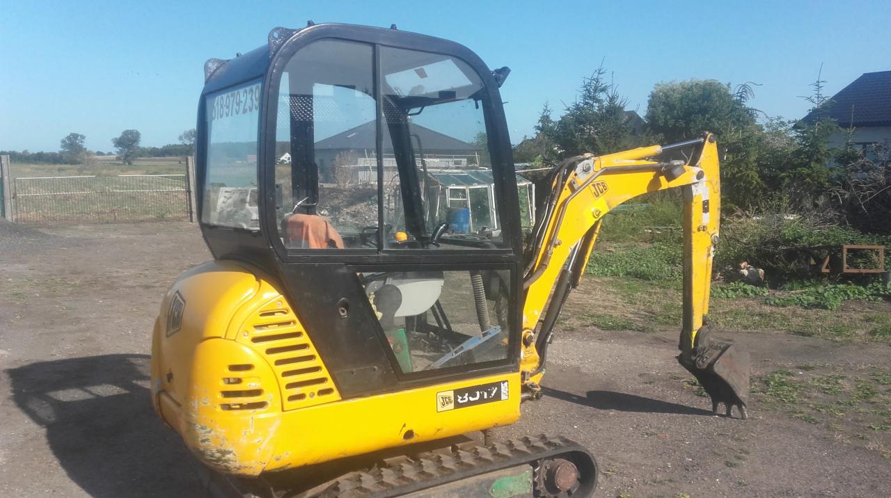 Żółta mini koparka JCB na gąsienicach, z łyżką lekko zanurzoną w ziemi, widoczna kabina operatora i otoczenie z zielenią oraz budynkiem w tle.