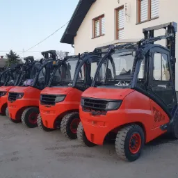 Szereg nowych, pomarańczowych wózków widłowych Linde, ustawionych przed budynkiem z anteną satelitarną, sugerujący gotowość do sprzedaży lub wynajmu.