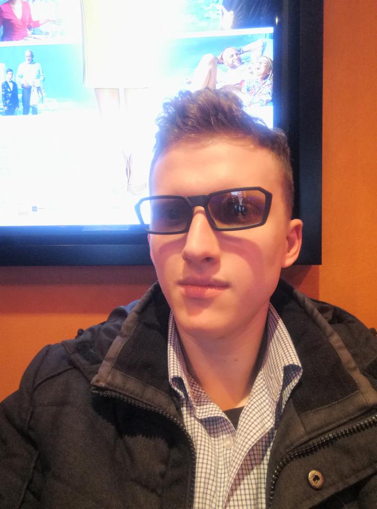 Młody mężczyzna w okularach 3D pozuje na tle ściany i ekranu z filmem, ubrany w kurtkę i koszulę w kratę.