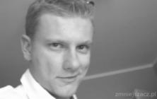 Portret młodego mężczyzny z krótkimi włosami, uśmiechniętego, w czarno-białej tonacji, z widocznym znakiem wodnym w prawym dolnym rogu.