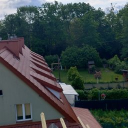 EKOMA SPORT - Czerwona dachówka ceramiczna na skośnym dachu domu jednorodzinnego z widocznymi kominami i oknami dachowymi, w tle zieleń drzew i zabudowa ogrodowa.