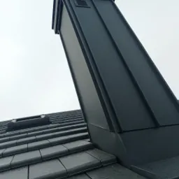 Czarny komin wentylacyjny z blachy na dachu pokrytym dach&oacute;wką ceramiczną, widoczny fragment okna dachowego. Ujęcie z dołu, pochmurne niebo w tle.