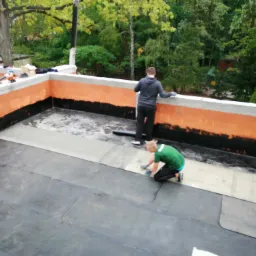 Prace na dachu płaskim: dwóch mężczyzn w trakcie układania papy termozgrzewalnej, jeden klęczy i dociska, drugi stoi przy murku z cegły pomalowanego od dołu na czarno. W tle drzewa.