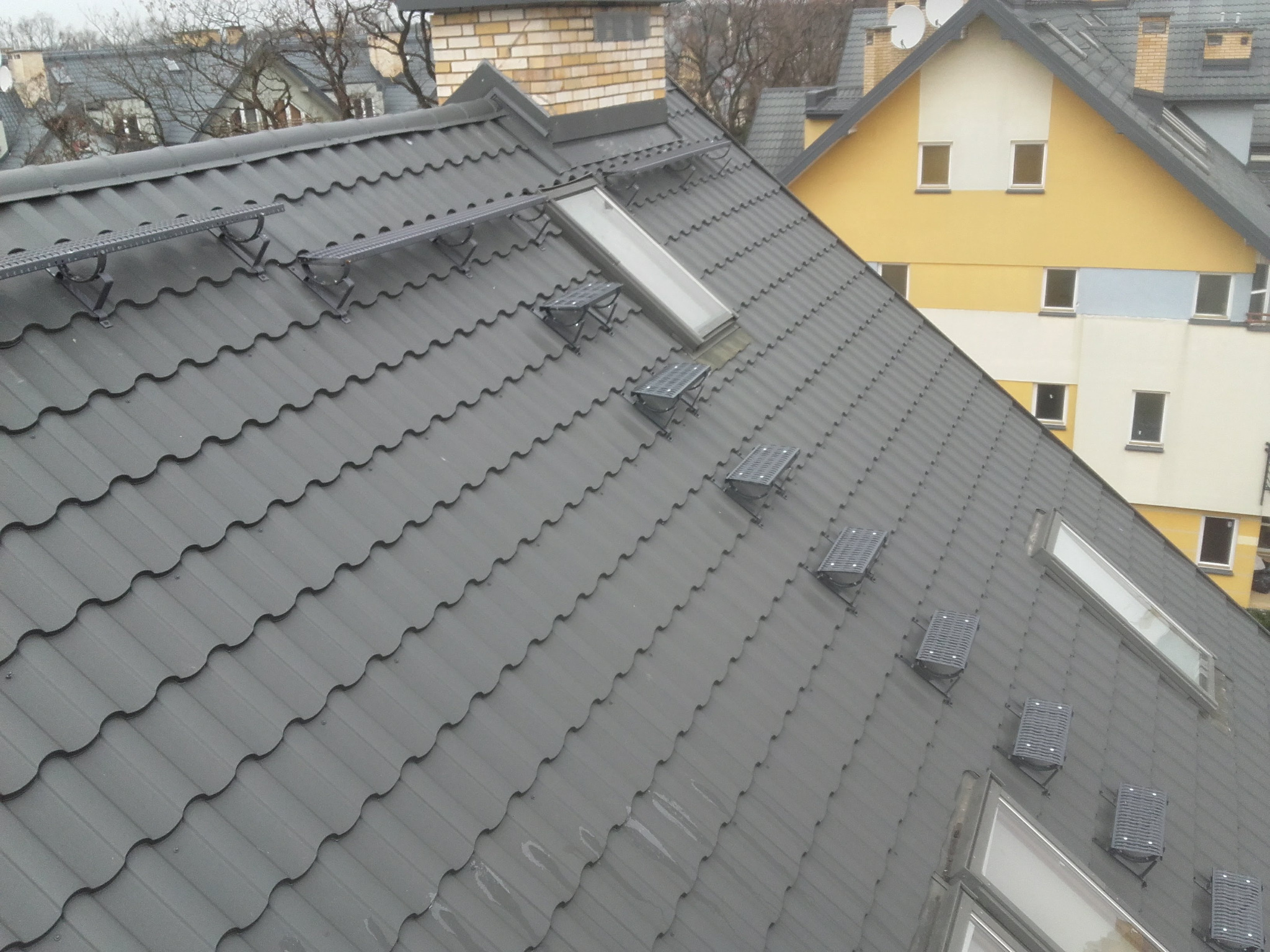 Szary dach z dachówki modułowej z zamontowanymi oknami dachowymi i elementami montażowymi. W tle budynek.