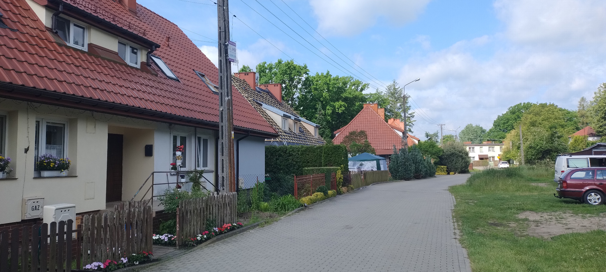 Ulica z domami w zabudowie szeregowej. Na jednym z domów widoczny remont dachu, odsłonięta konstrukcja i przygotowanie do położenia nowego pokrycia.