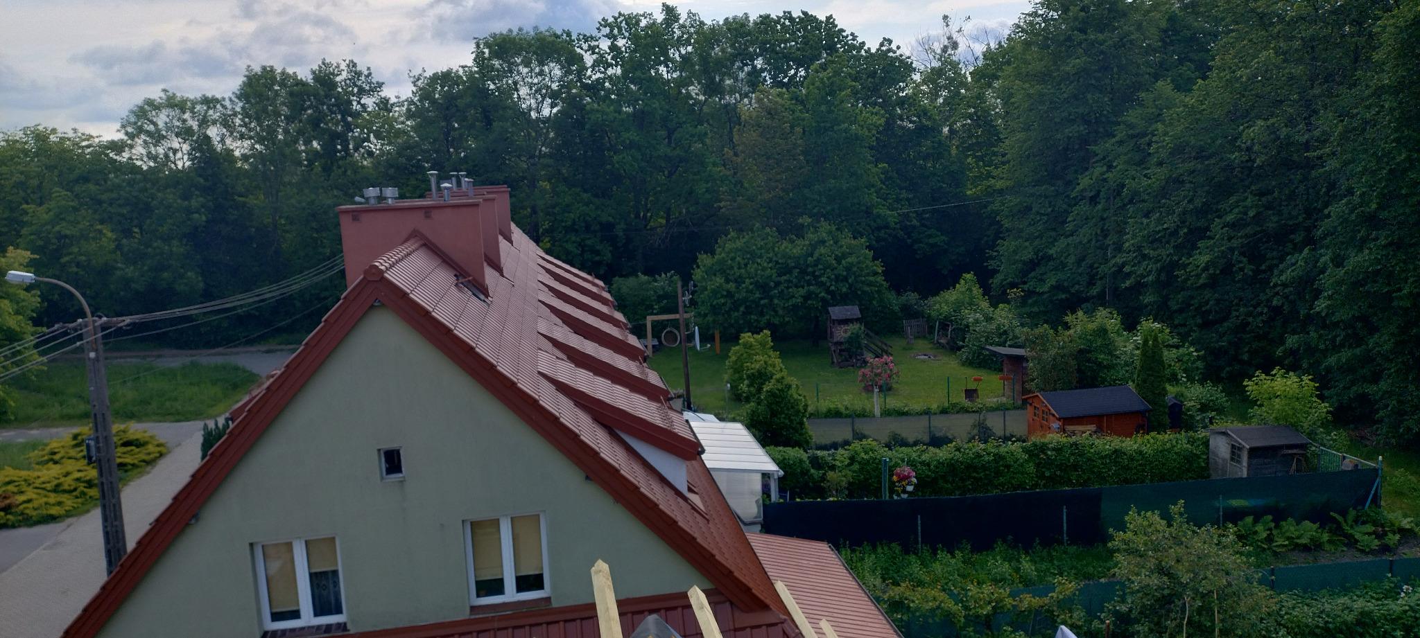 Czerwona dachówka ceramiczna na skośnym dachu domu jednorodzinnego z widocznymi kominami i oknami dachowymi, w tle zieleń drzew i zabudowa ogrodowa.