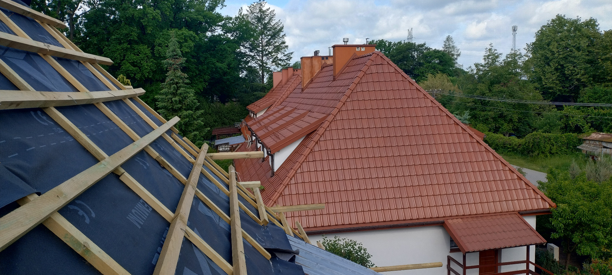 Częściowy widok dachu w trakcie budowy z drewnianą konstrukcją i membraną, w tle gotowy dach z czerwonej dachówki ceramicznej i zieleń drzew.