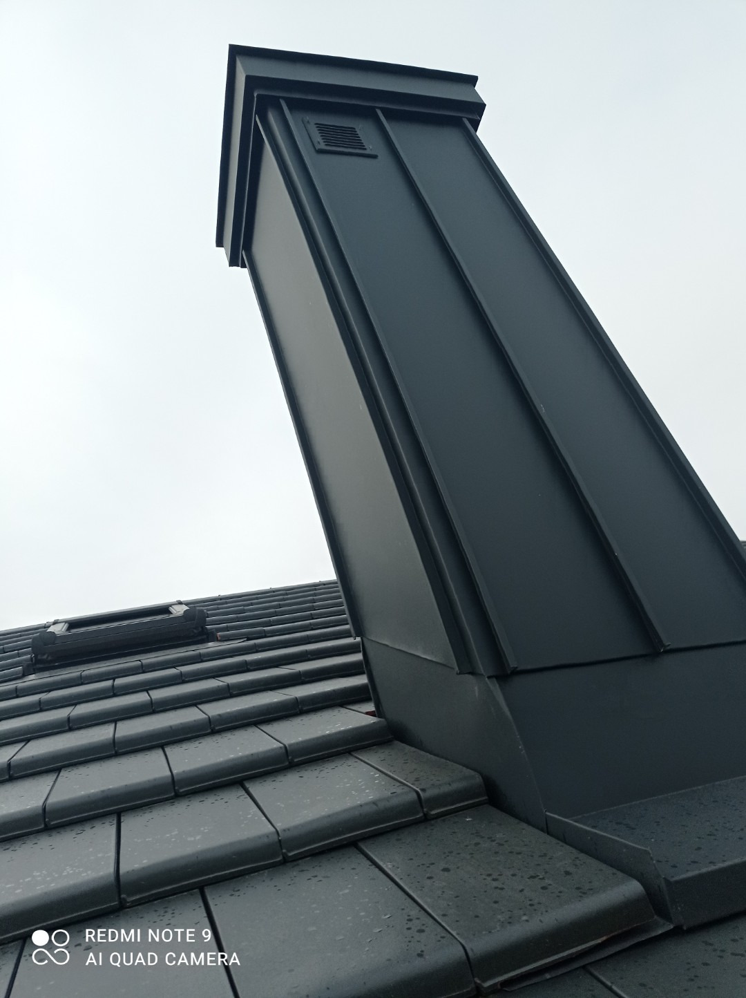 Czarny komin wentylacyjny z blachy na dachu pokrytym dachówką ceramiczną, widoczny fragment okna dachowego. Ujęcie z dołu, pochmurne niebo w tle.