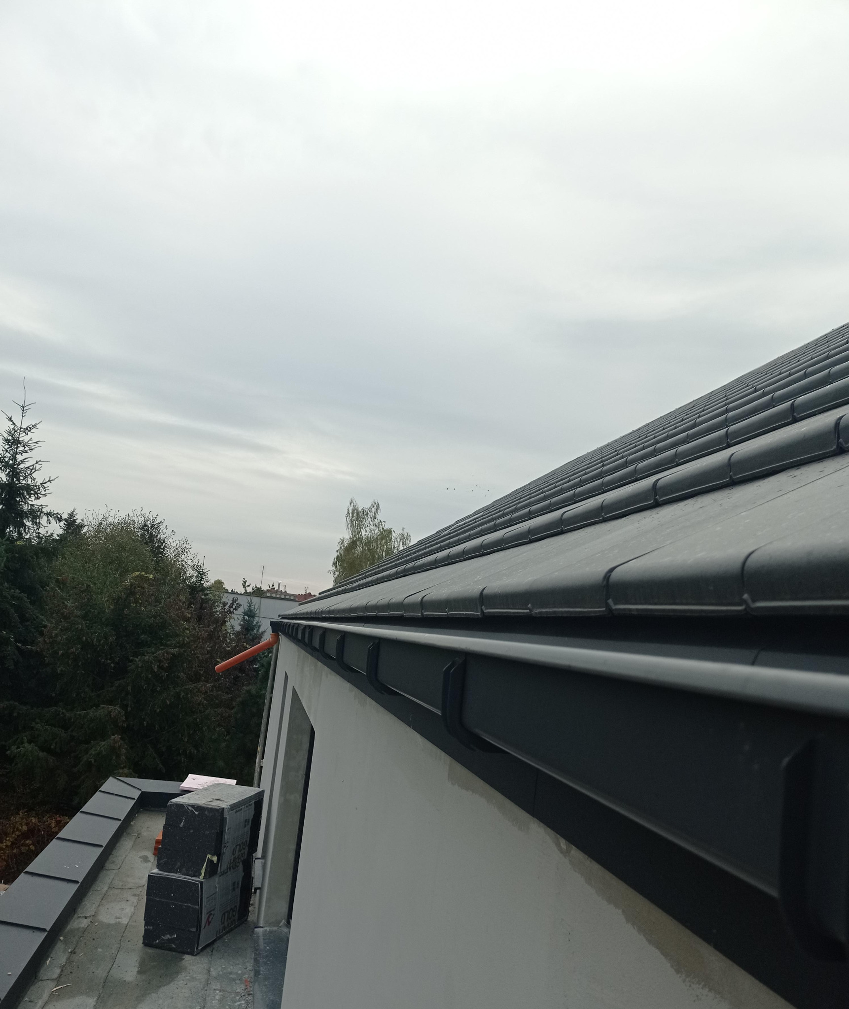 Czarny dach z dachówki ceramicznej, widok z dołu pod kątem, rynna wzdłuż ściany budynku, drzewa w tle, pochmurne niebo.
