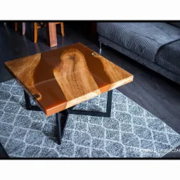 Kwadratowy stolik kawowy z blatem z drewna i żywicy epoksydowej w odcieniach brązu, na czarnych, metalowych nogach, ustawiony na szarym dywanie w geometryczny wzór. W tle widoczny fragment szarej...