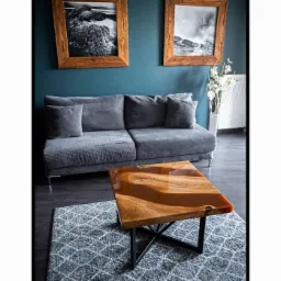 Stolik kawowy z blatem z drewna i żywicy epoksydowej na metalowej podstawie krzyżakowej, ustawiony na wzorzystym dywanie w salonie z szarą sofą i obrazami w drewnianych ramach na ścianie.