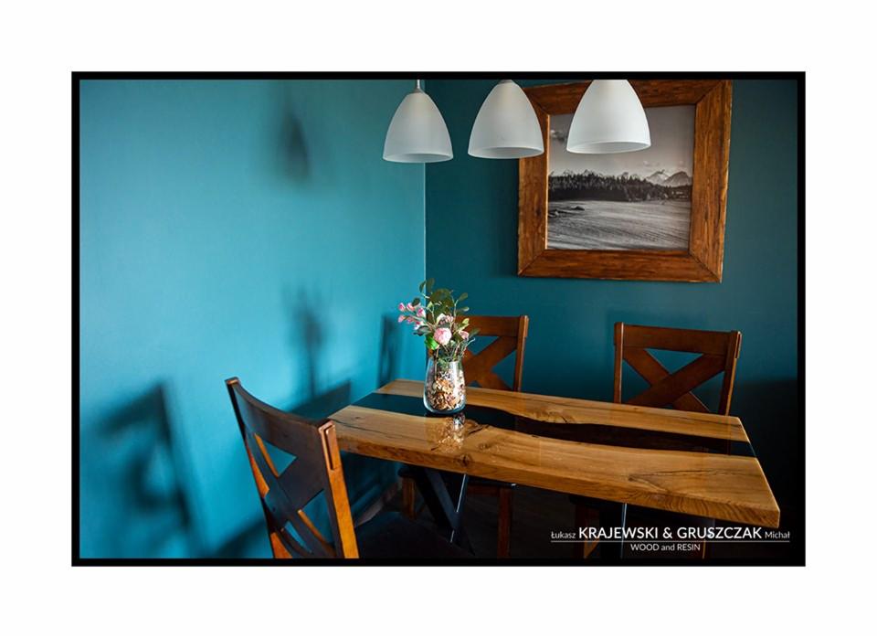 Stół z blatem z drewna i żywicy epoksydowej na metalowych nogach, ustawiony w rogu pokoju z turkusowymi ścianami, nad stołem wiszą trzy białe lampy, a na ścianie wisi obraz w drewnianej ramie.